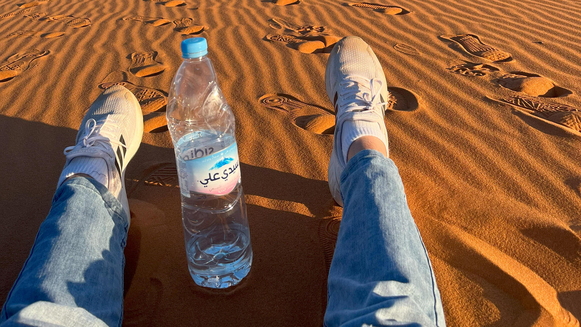 Beine im Wüstensand in Marokko mit Wasserflasche mit arabischer Schrift