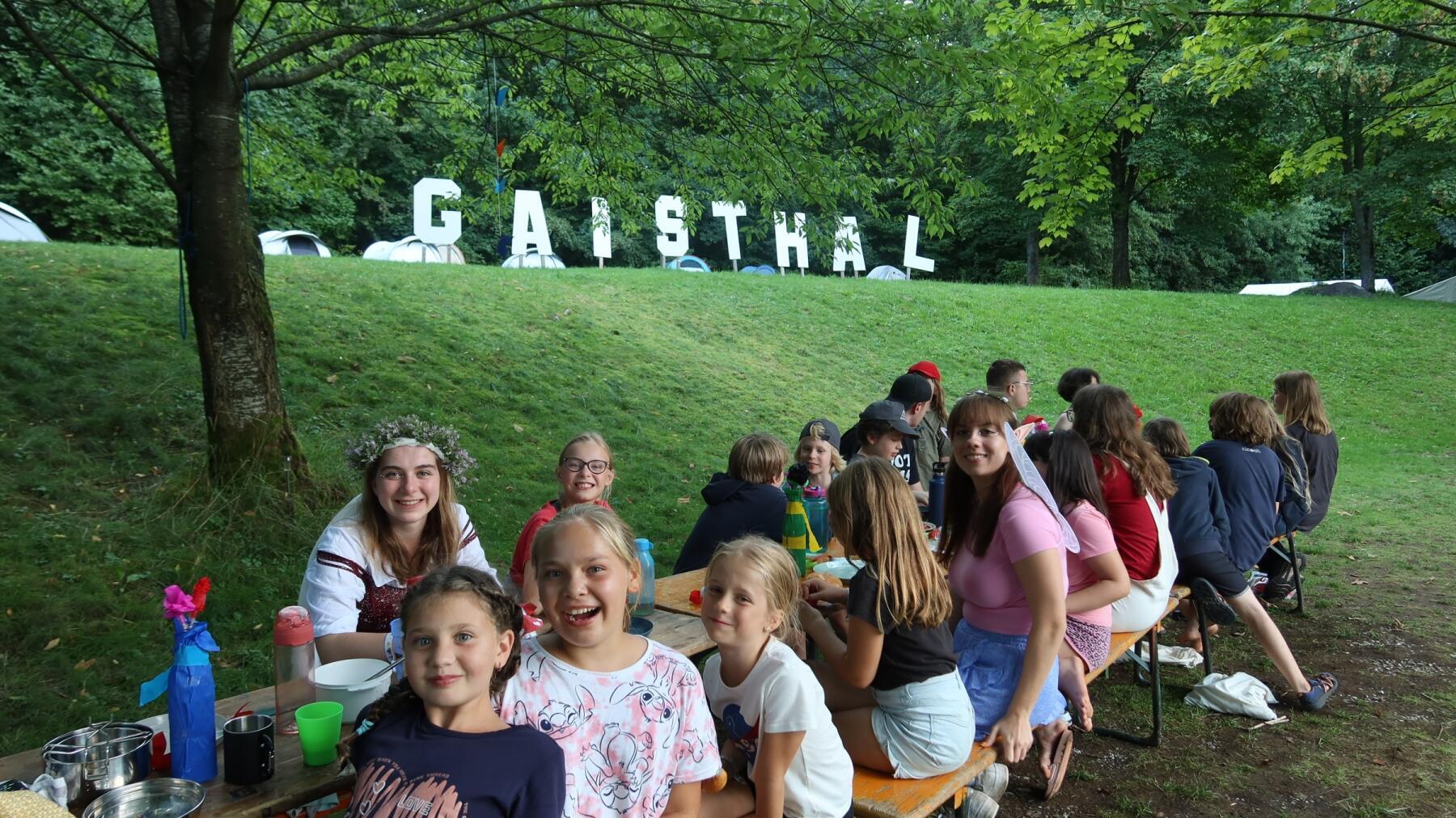 Kinder sitzen mit Betreuenden auf Bierbänken auf einer Wiese