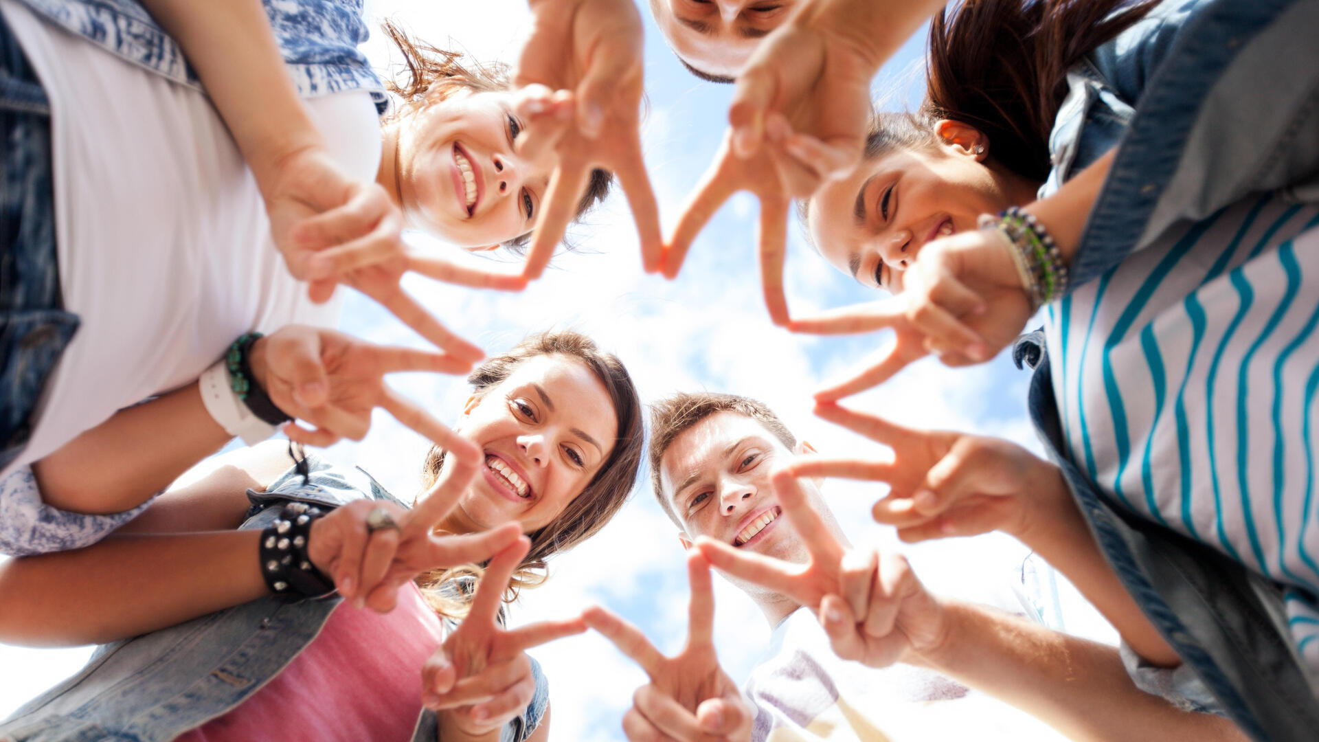 Fünf junge lachende Menschen im Kreis gucken mittig nach unten und geben ein Friedenszeichen mit ihren Fingern